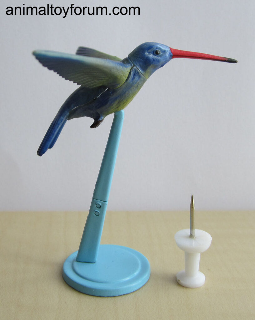 hummingbird figuring in a flying pose on a blue base, with a pinboard pin for scale.